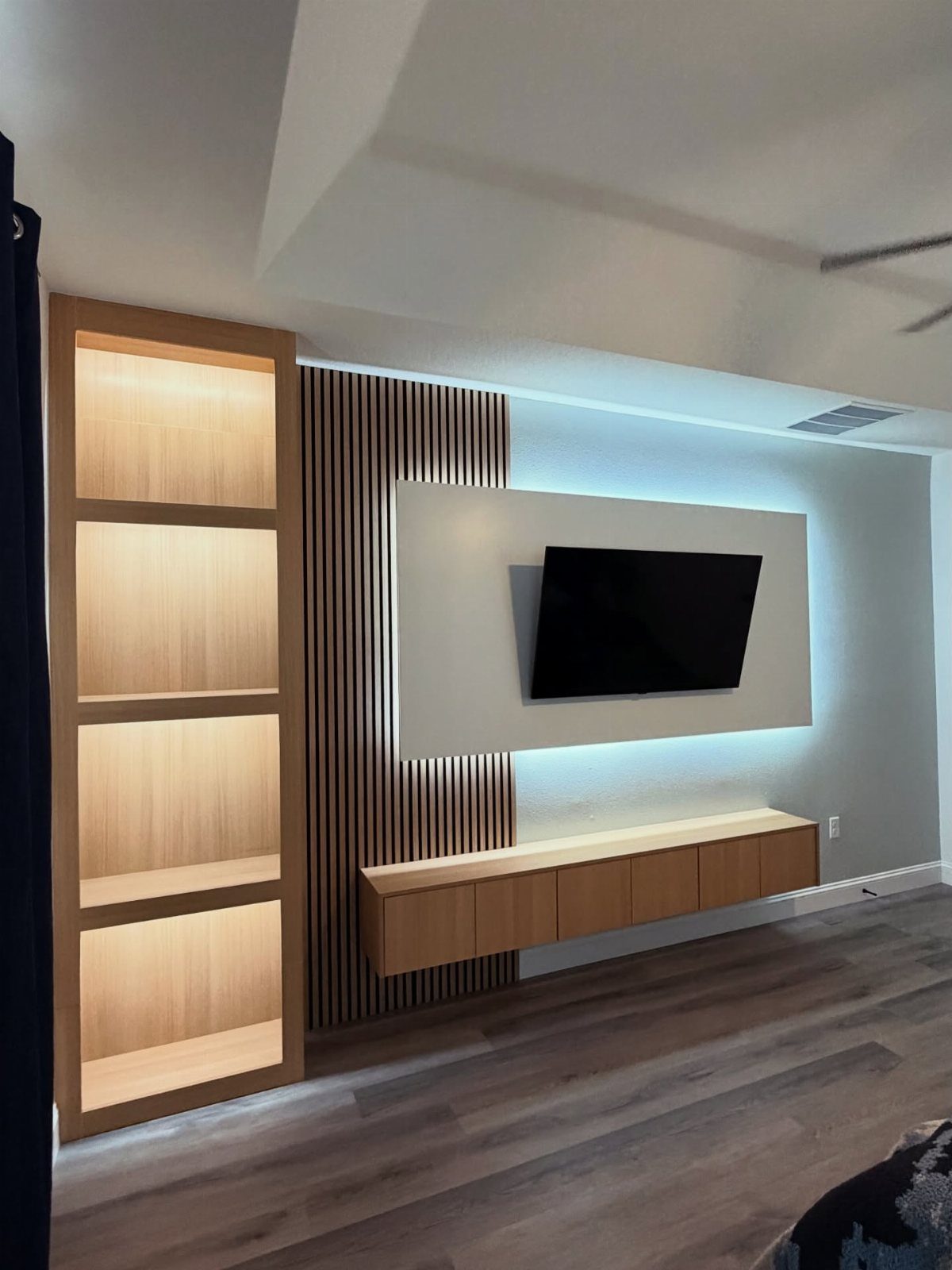 A television mounted on a backlit white panel next to wood slats and lit shelving.