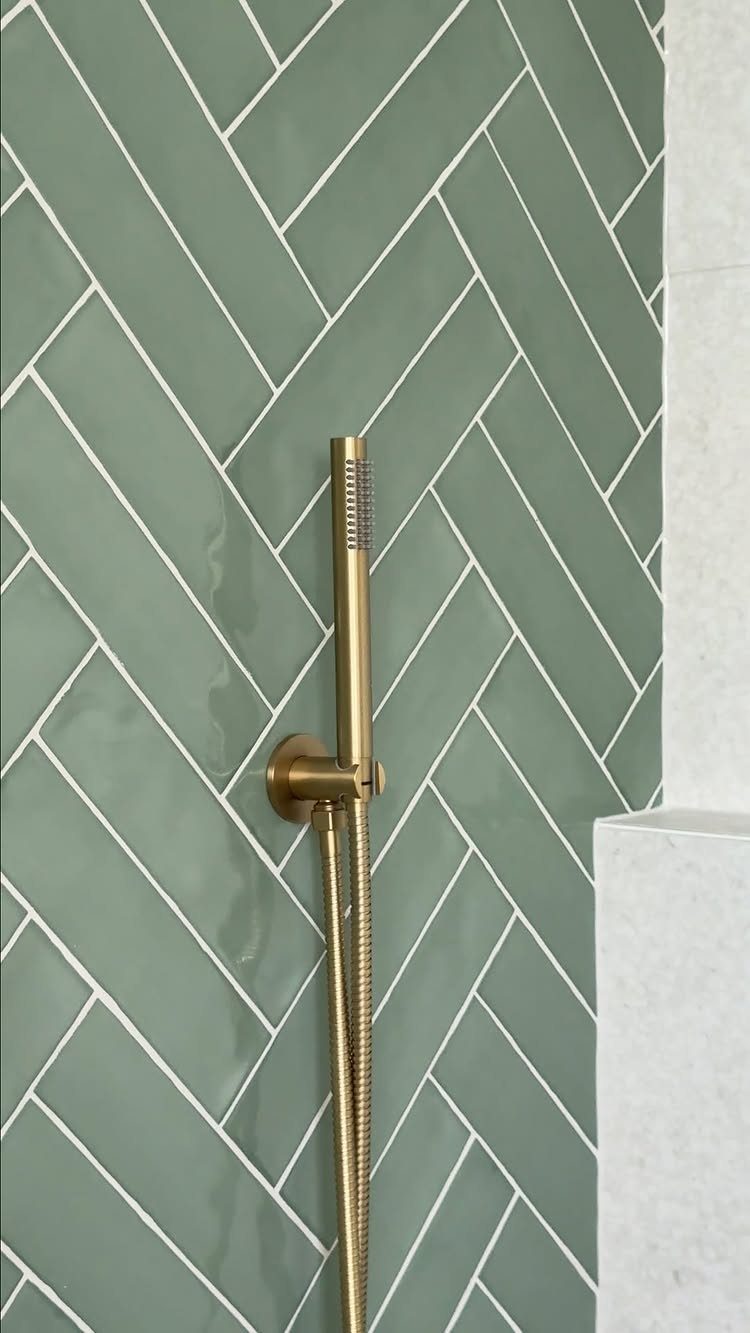 Sage green herringbone tiles with a gold handheld showerhead mounted on the wall.