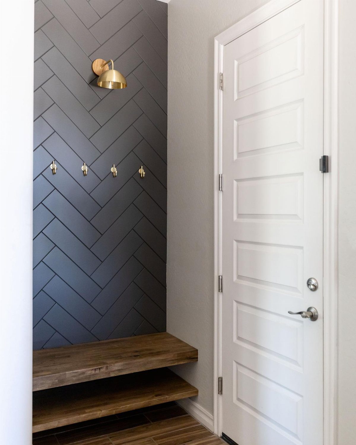 Dark gray herringbone pattern wall trim with gold hooks and a sconce in an entryway.