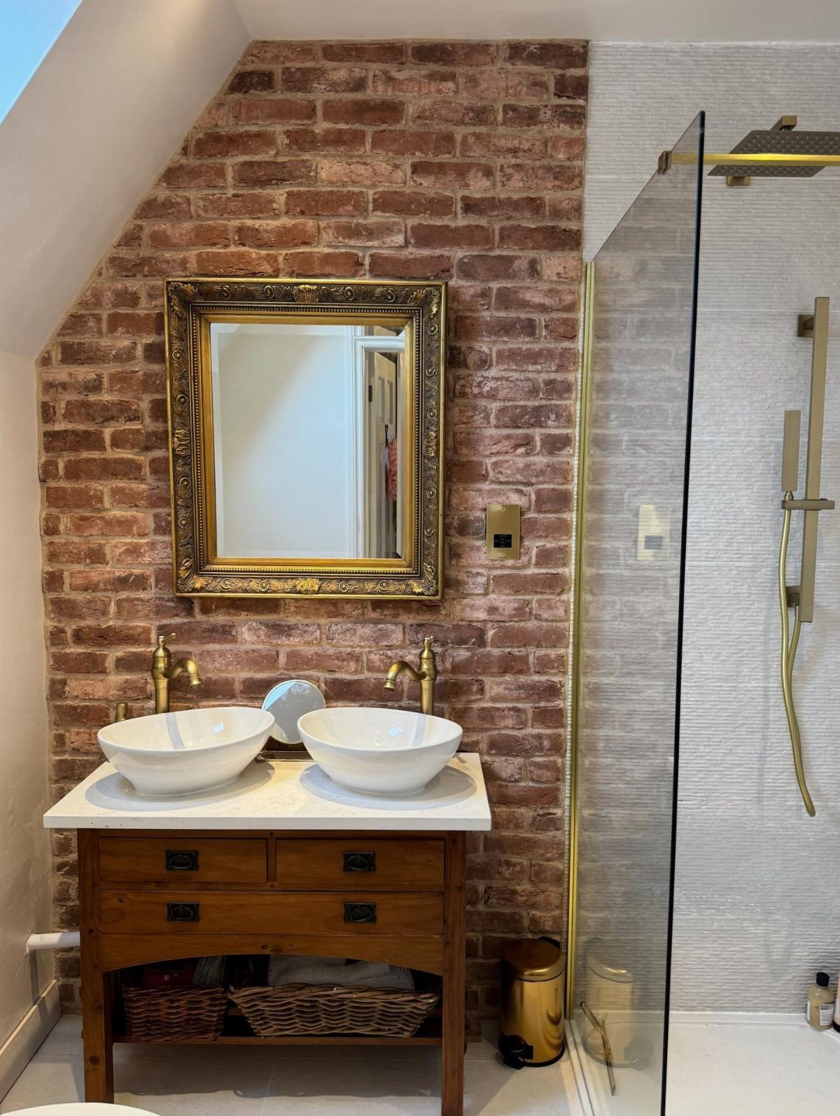 Red brick accent wall in a bathroom with a wooden vanity and gold fixtures.