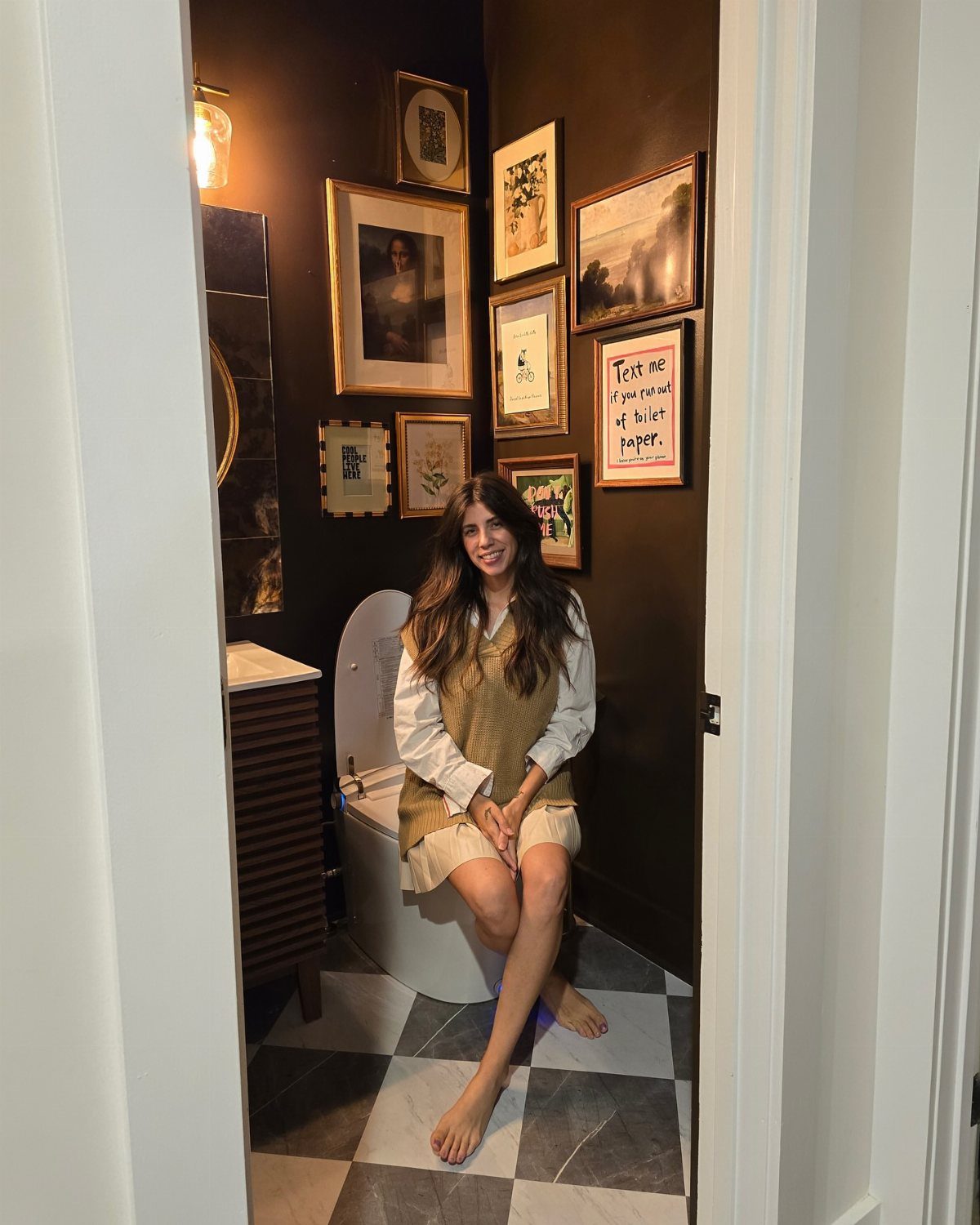 Woman sitting on a toilet in a bathroom with a dark gallery wall.