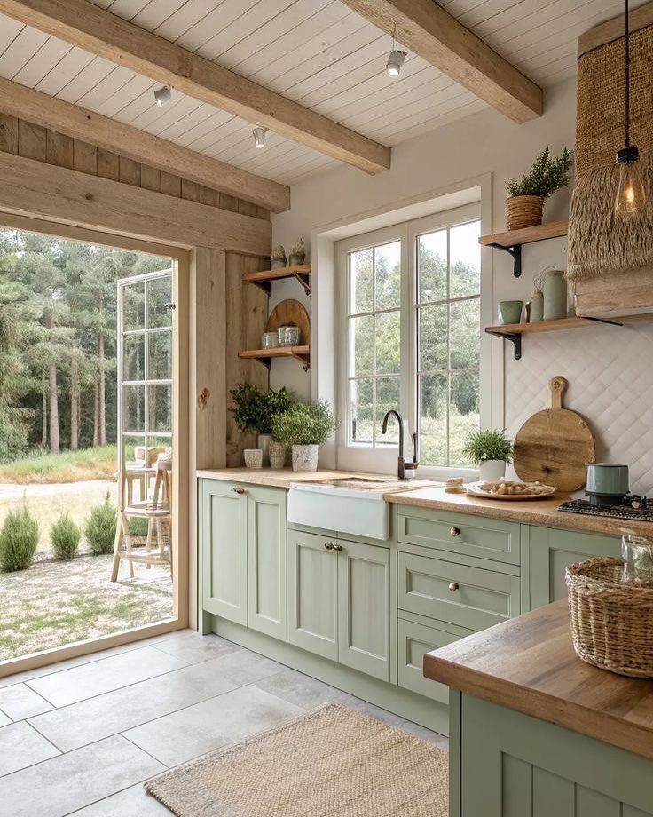 Sage green kitchen cabinets with open wooden shelves and a white farmhouse sink near a window.