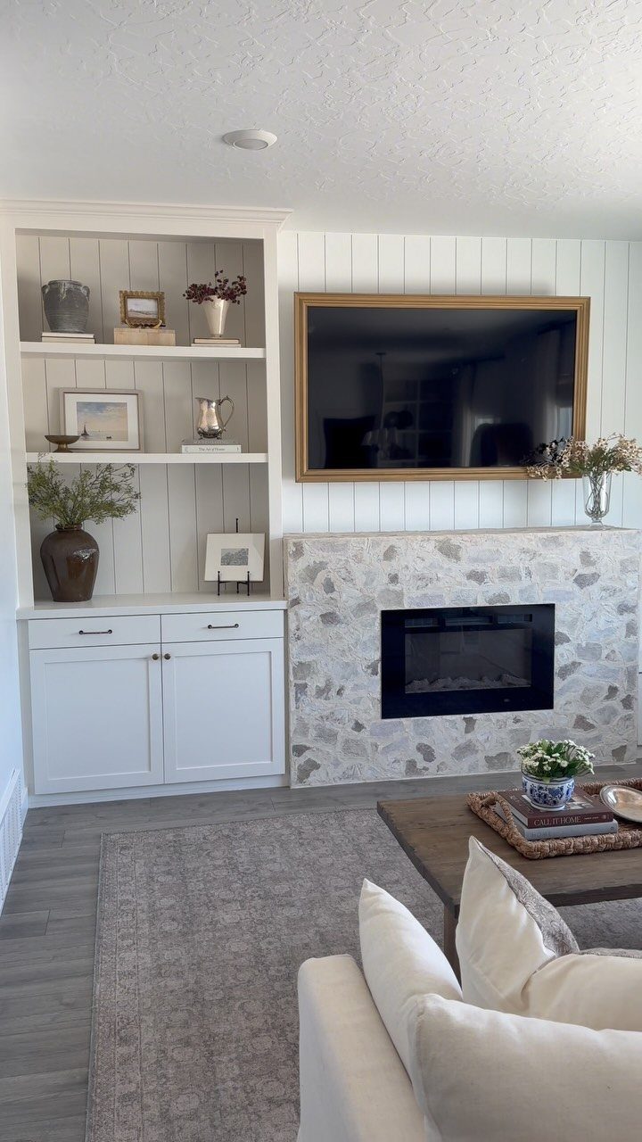 Grey flooring in a living room with a white bookshelf, stone fireplace, and gold-framed television.