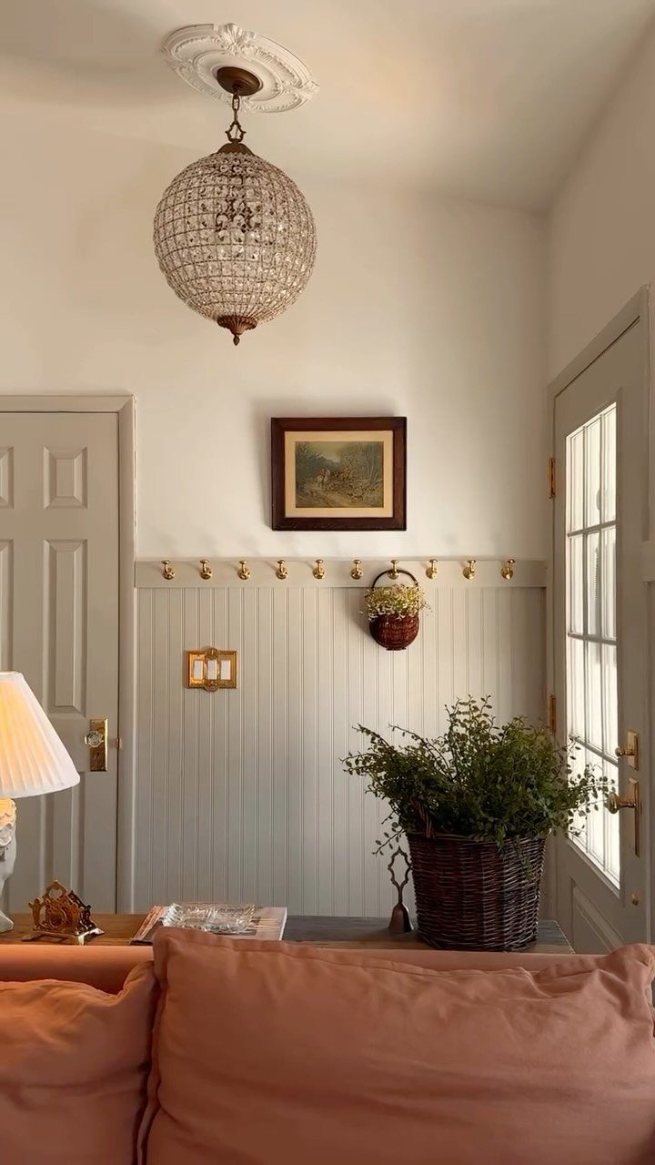 Light grey beadboard wainscoting on a white wall with brass hooks and a crystal chandelier.