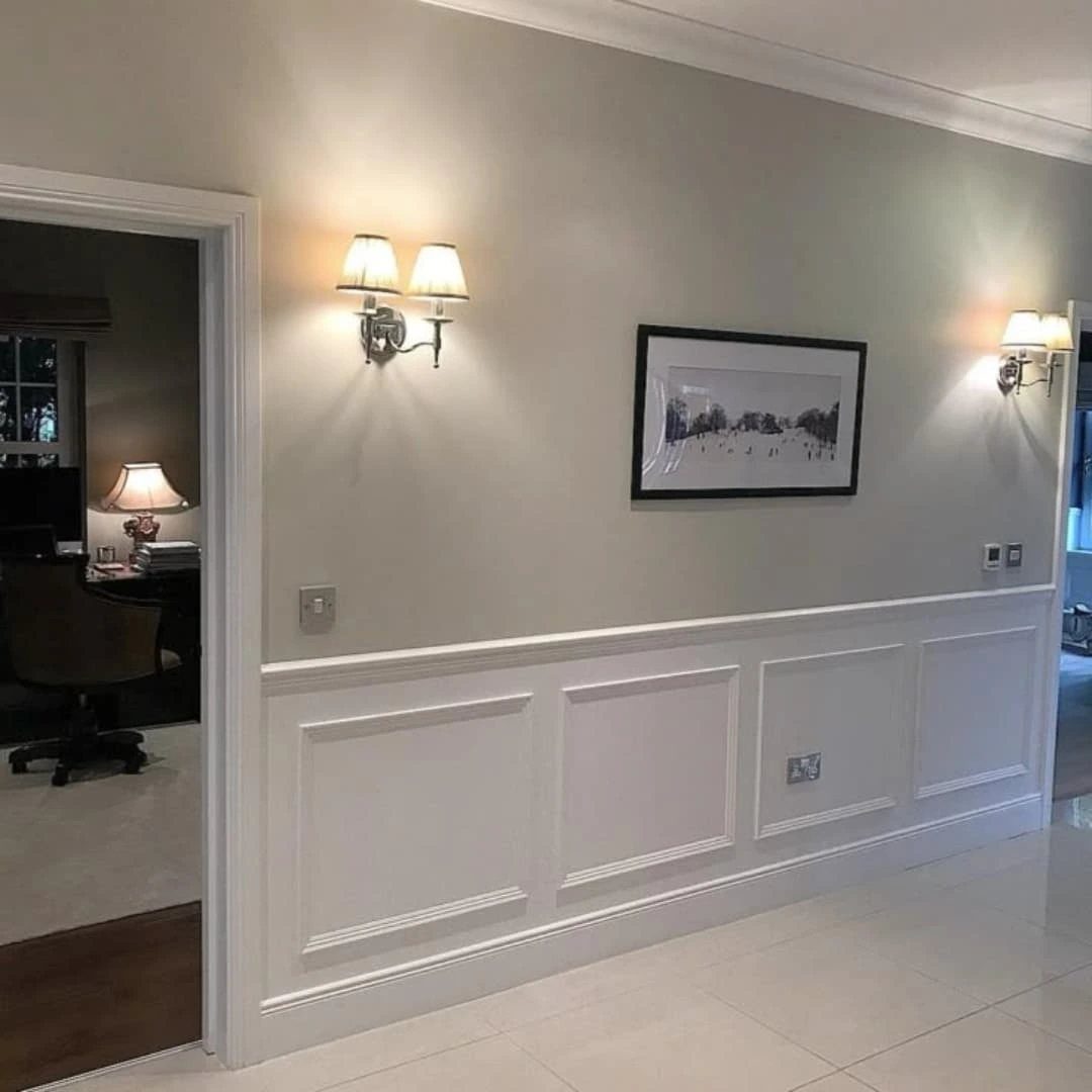 White wainscoting with rectangular molding panels on a light grey wall with wall sconces.