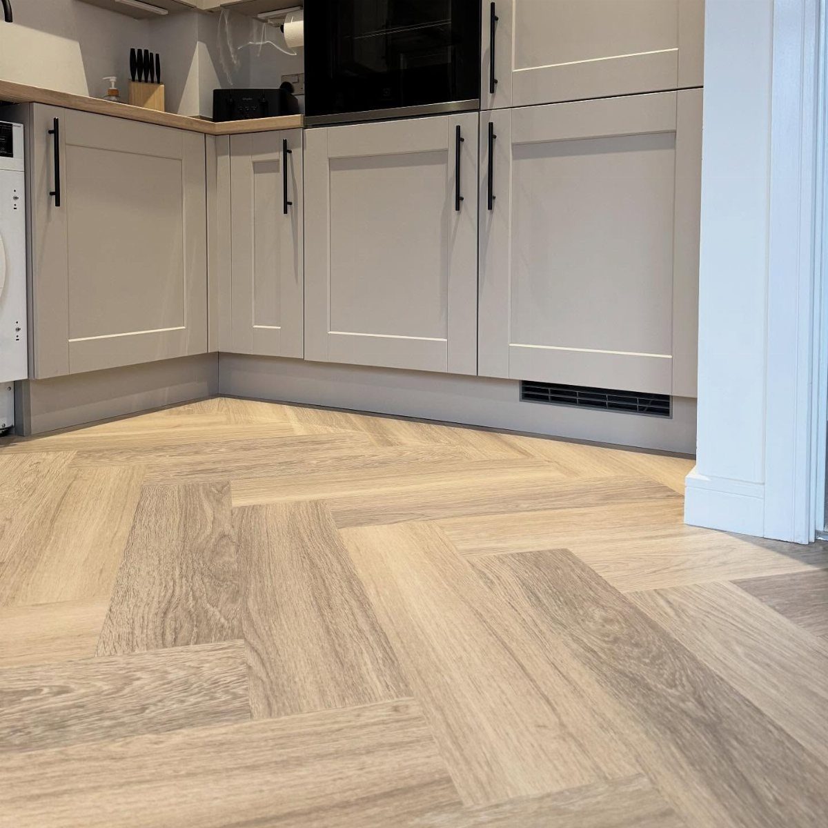 Light wood-look planks arranged in a herringbone pattern on a kitchen floor near grey cabinets.