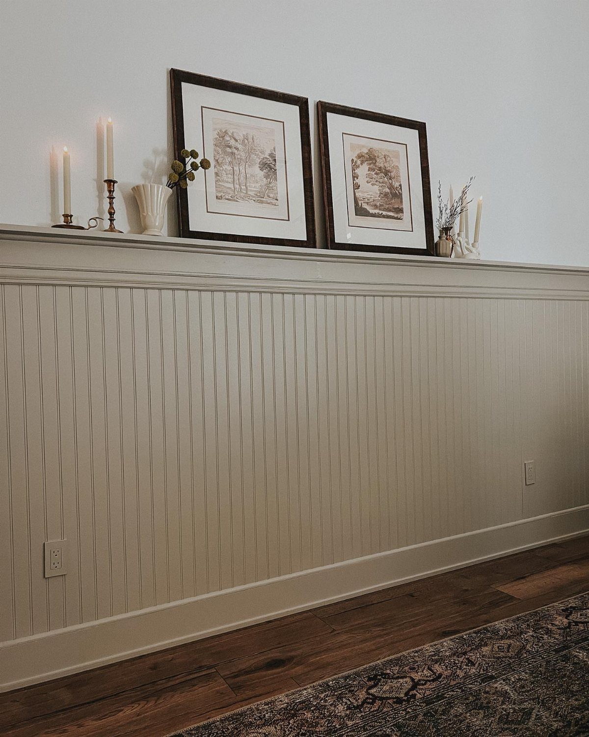 Vertical cream beadboard wainscoting on a wall with framed art and candles on a shelf.