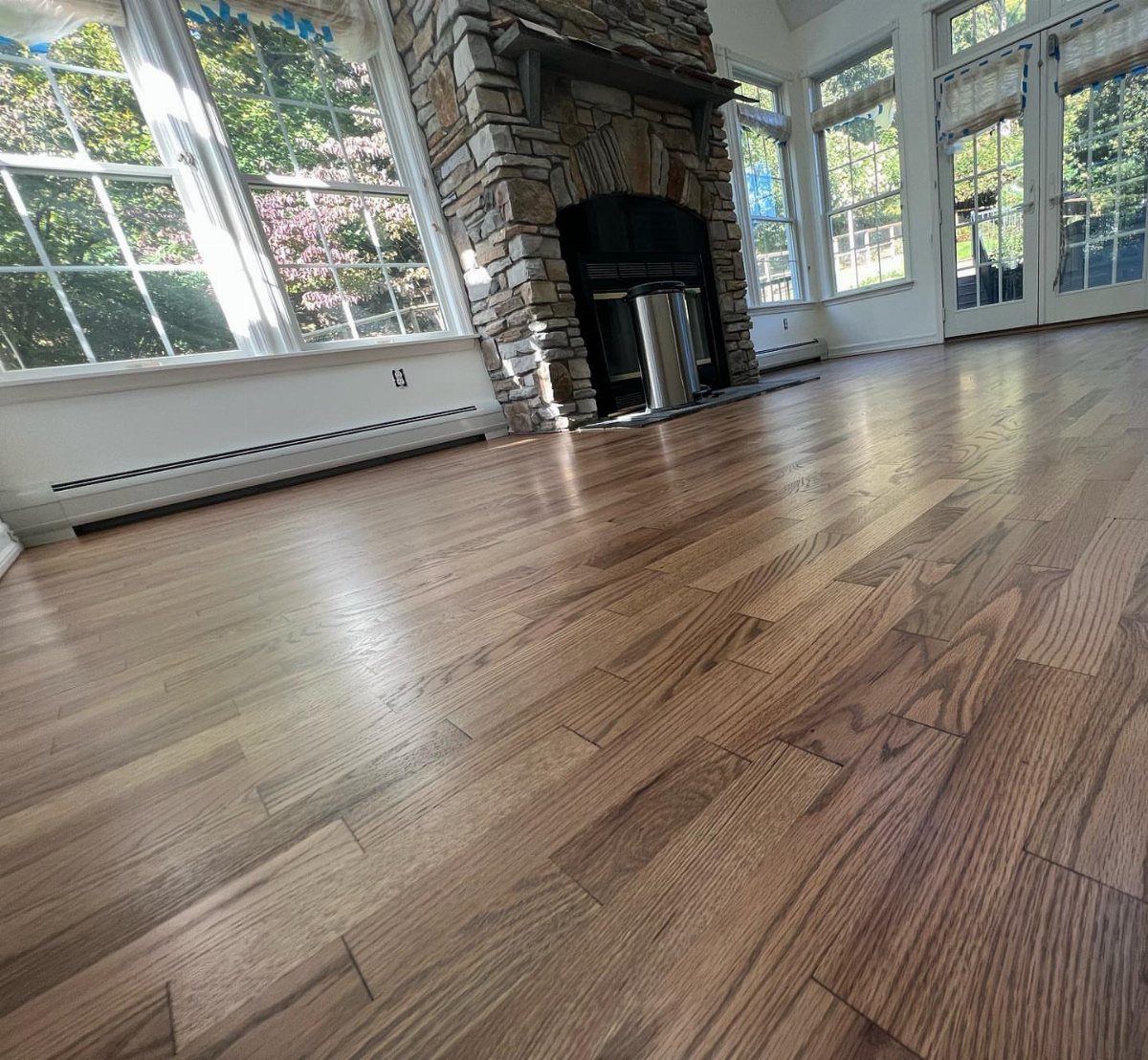 Polished hardwood floors with a light brown grain stretching across a bright living room with a stone fireplace.