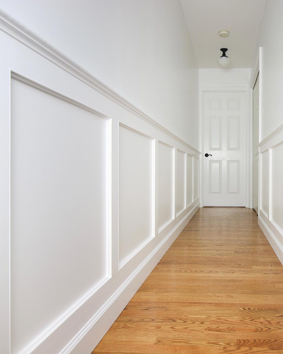 White board-and-batten wainscoting installed along a bright hallway with light wood floors.