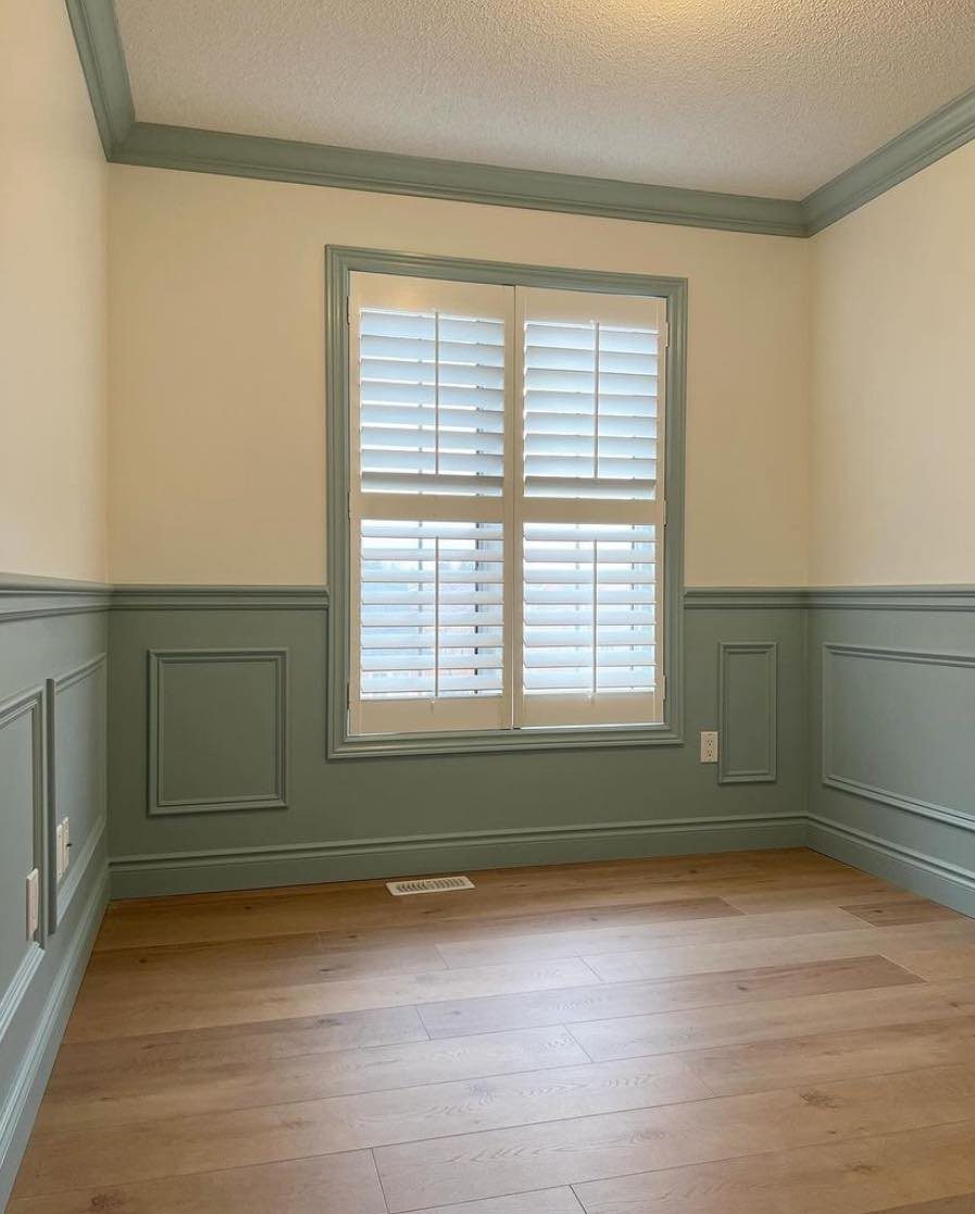 Sage green paneled wainscoting paired with neutral cream walls and a window with white shutters.