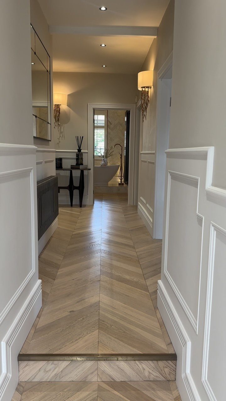 White decorative wainscoting lines a hallway with light wood herringbone floors and neutral walls.