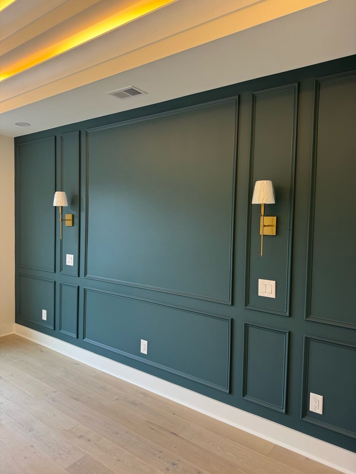 Dark teal wainscoting with rectangular molding and gold wall sconces on a large interior wall.