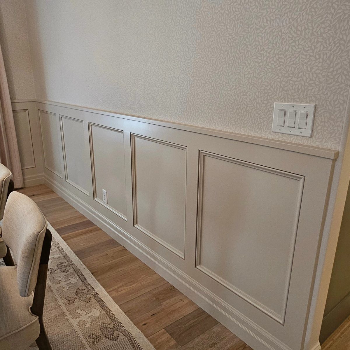 Light gray painted wainscoting panels installed on a wall beneath patterned wallpaper in a dining room.