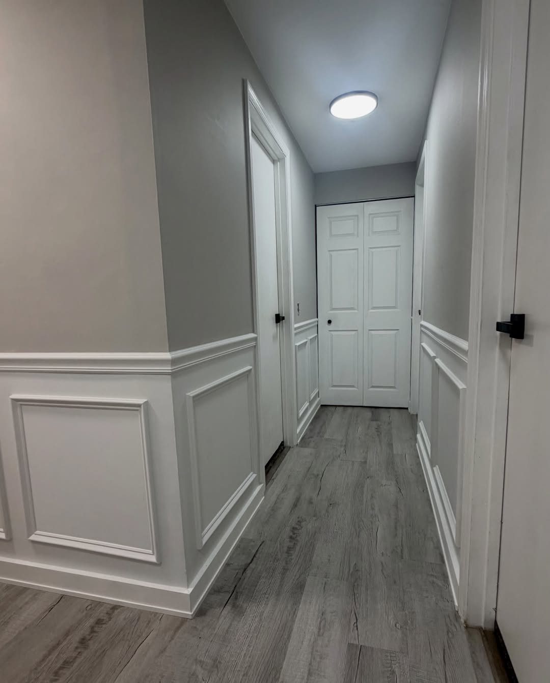 A hallway with grey walls decorated with white picture frame wainscoting and light wood flooring.