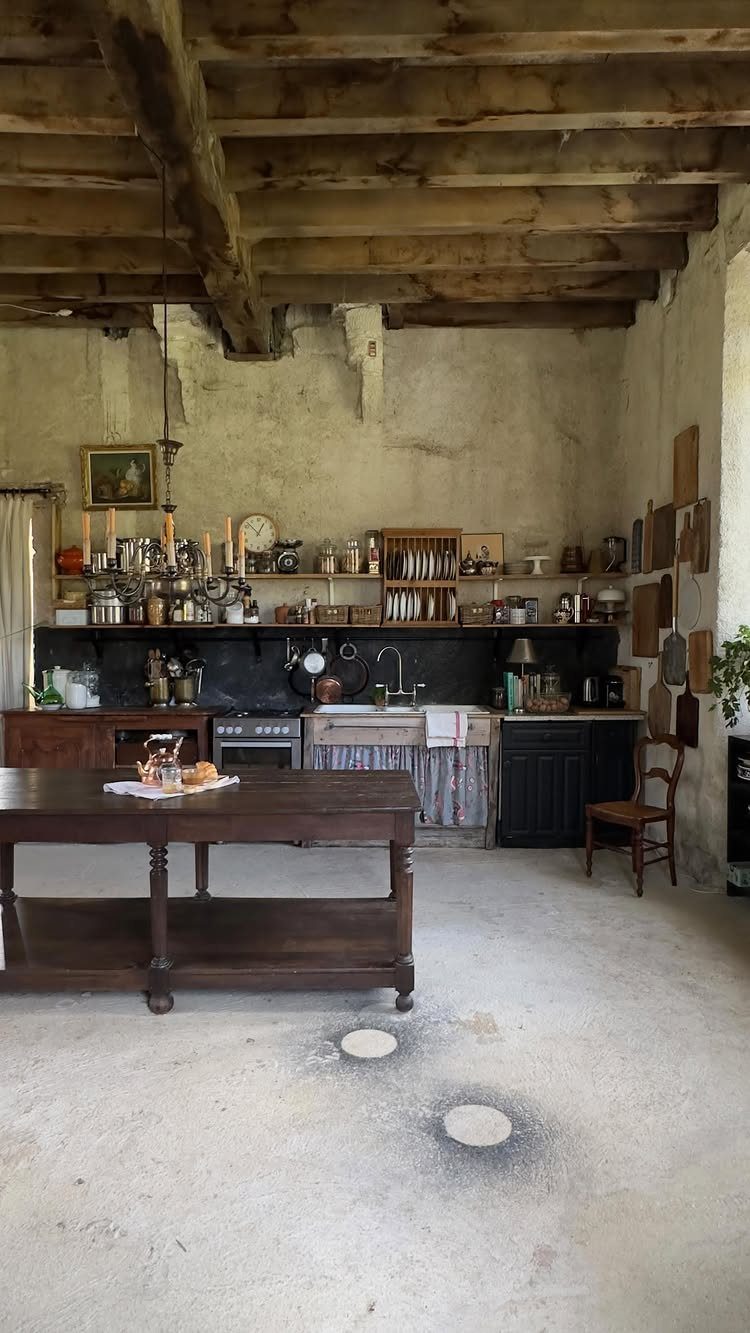 Rustic kitchen featuring wooden ceiling beams, stone walls, a large wooden island, and a weathered concrete floor.
