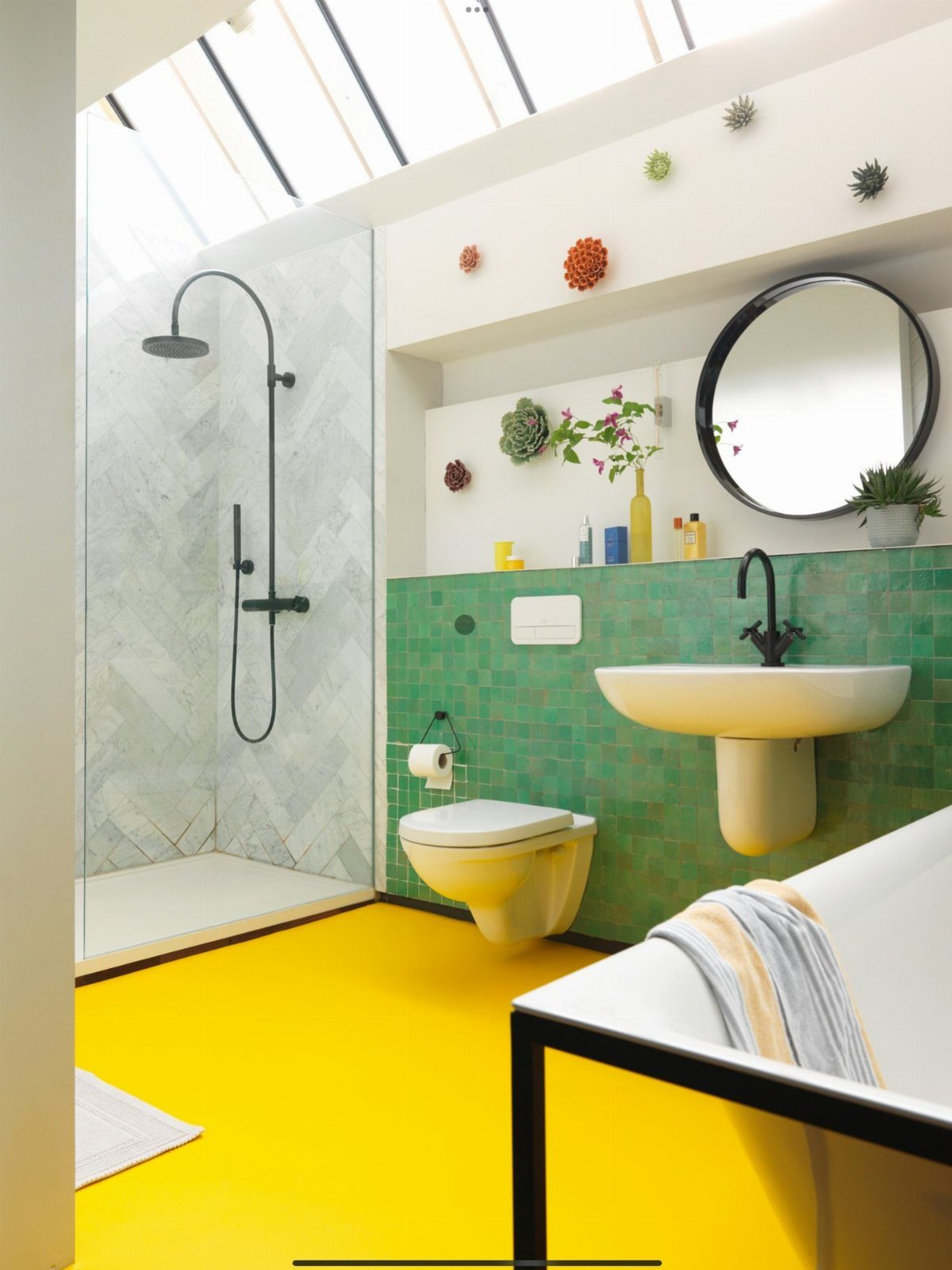 A bright bathroom with bold yellow flooring, green wall tiles, and a glass shower enclosure.