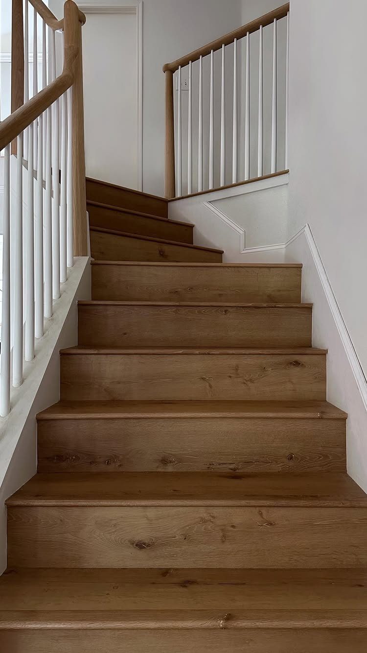 Light brown wooden stairs with white spindles and a wooden handrail in a bright room.
