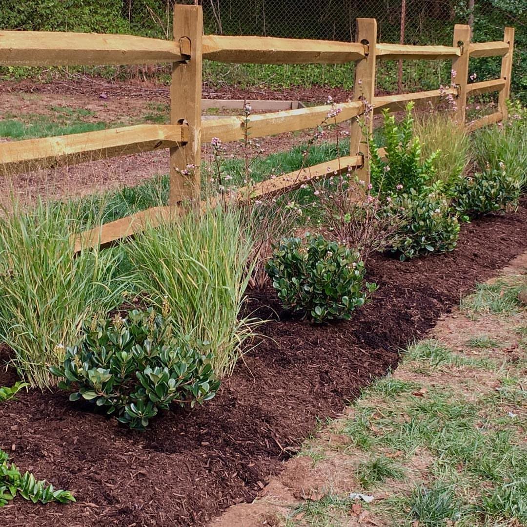 A rustic split-rail wooden fence bordering a garden bed with green plants and dark mulch.