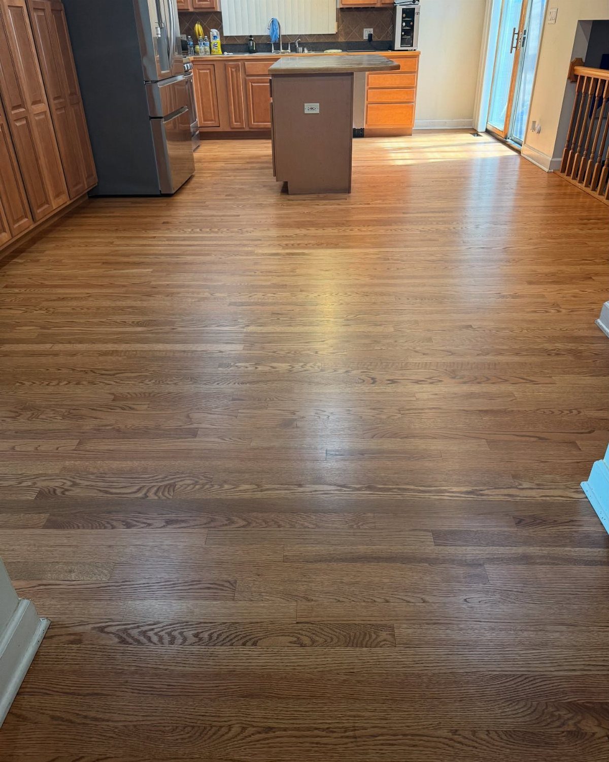 Medium brown wood plank flooring in a kitchen with wooden cabinets and a stainless steel refrigerator.