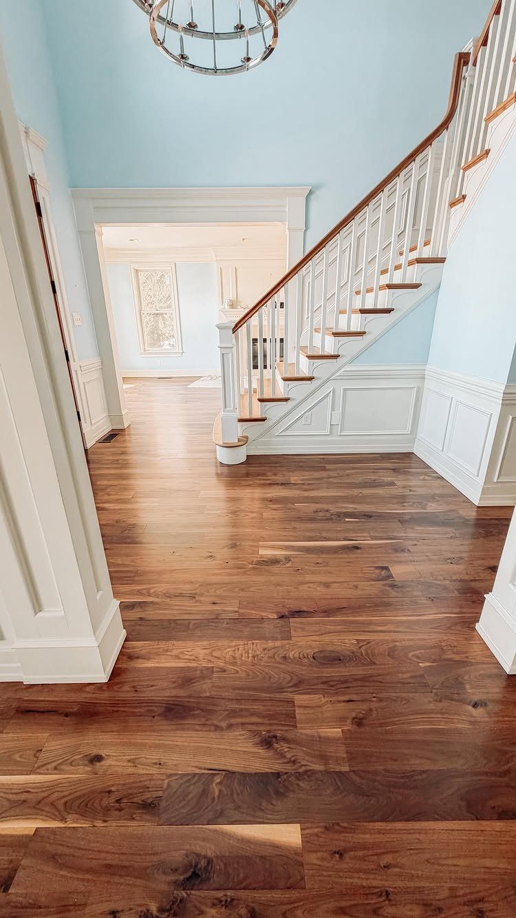 Medium brown wood plank flooring in a hallway with white wainscoting and a staircase.