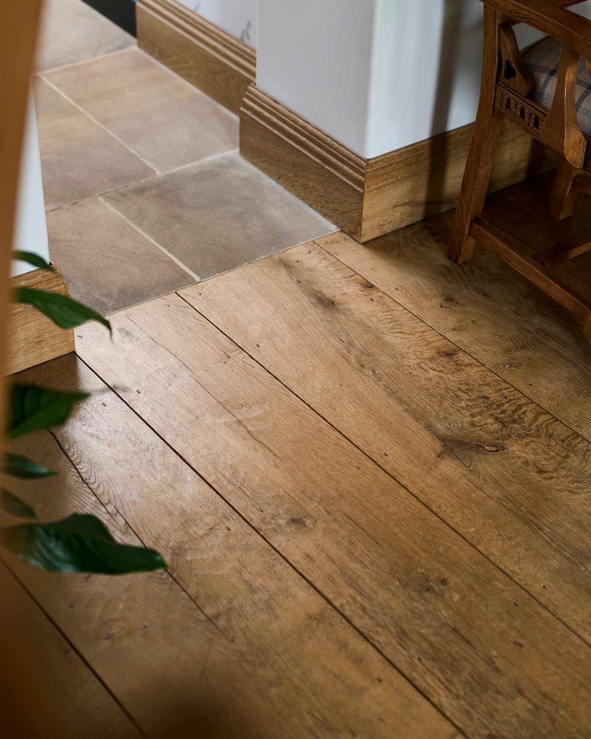 Light brown reclaimed wooden floor planks meeting grey tiles and a white wall with baseboards.